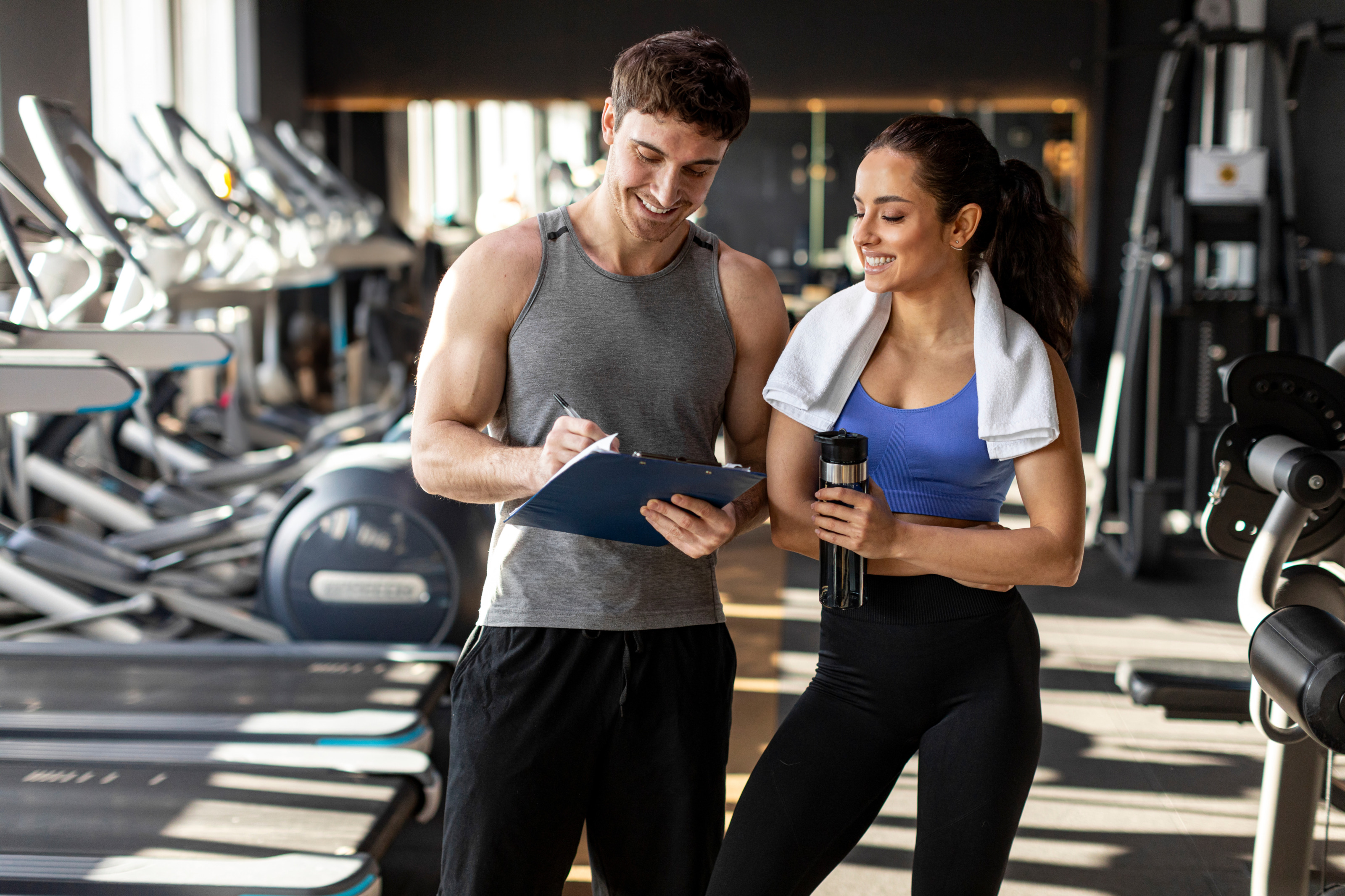 Zwei Personen in Sportkleidung stehen nebeneinander. Die linke Person notiert etwas auf einem Klemmbrett, auf das beide blicken. Die rechte Person hat ein Handtuch um den Hals. Im Hintergrund stehen verschiedene Sportgeräte