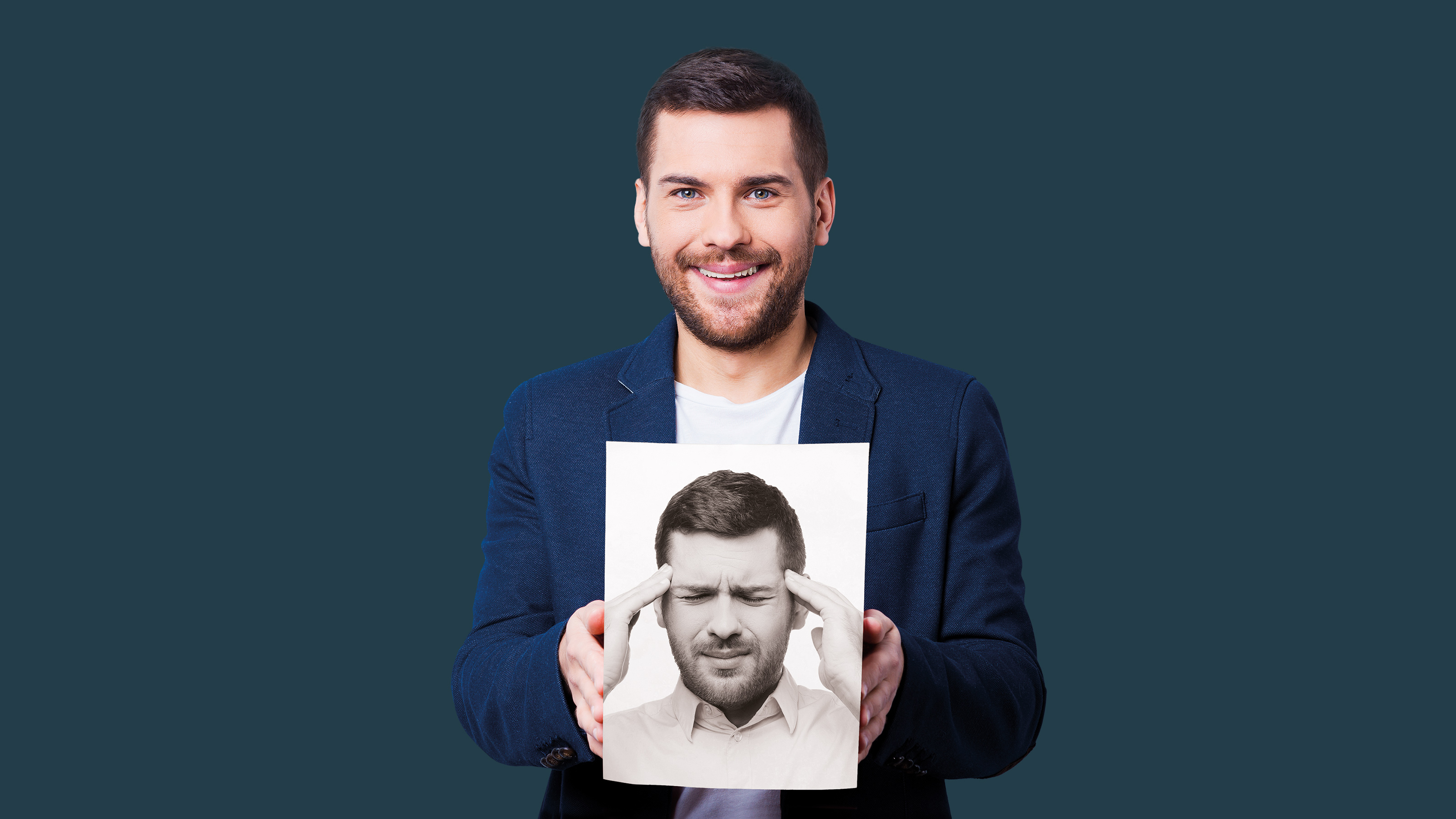 Junger Mann in formeller Kleidung, der vor einem dunkelblauen Hintergrund steht und ein Schwarz-Weiß-Bild von sich selbst hält, in dem er seinen Kopf berührt und die Augen vor Schmerz geschlossen hält.